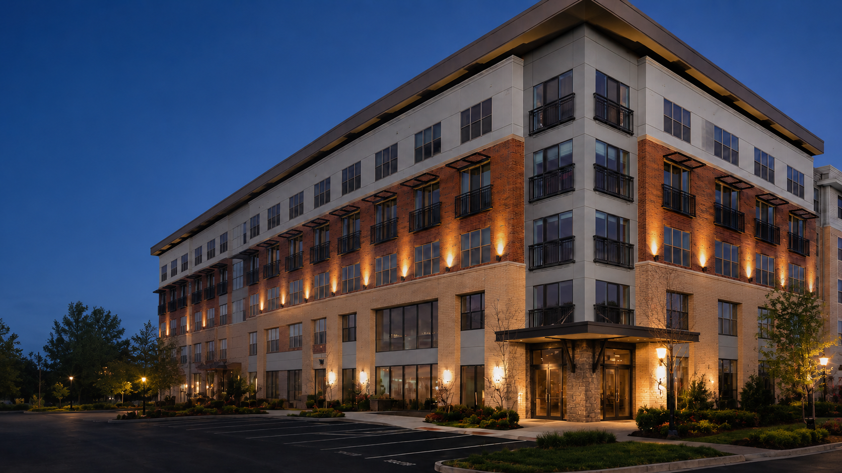Toscana - Passco Companies Modern four-story apartment building at dusk with brick and stone facade, warm exterior lights, and a front parking lot.